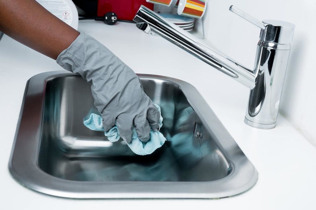 Gloved hand cleaning a stainless steel sink with a blue cloth.